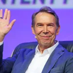 A smiling white man in a suit raising one hand.