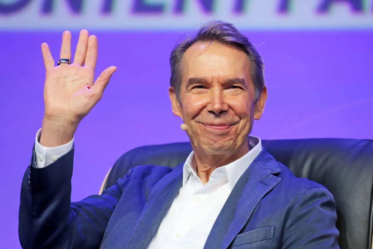 A smiling white man in a suit raising one hand.