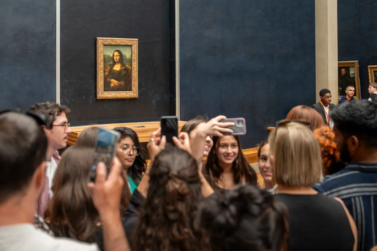 A crowd of people, some holding smartphones, stands before a painting of a woman with her arms in her lap.