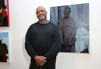 A Black man in a black sweater standing beside a painting of a Black man in similar clothes.