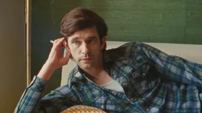 A white man lying on a couch with a cigarette in one hand. He wears a plaid shirt and has propped one hand under his head.
