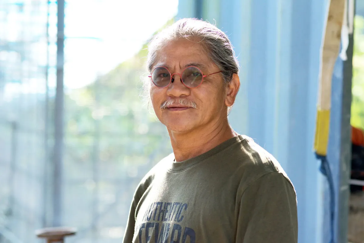 Portrait of a smiling Indigenous man with a salt-and-pepper mustache and round red-framed glasses. He stands in front of a window with light pouring in, wearing a T-shirt.