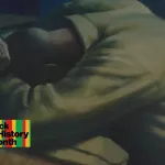 A painting of a Black man leaned over a table with his hands over his head.