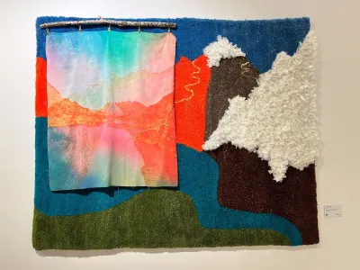 A textile work featuring a mountain range with a cloud passing across it. A hanging fabric covers part of the range.