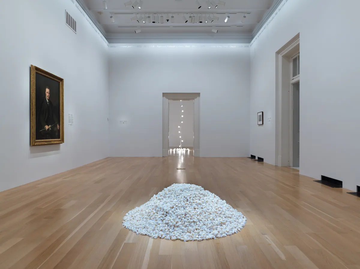 A gallery with a pile of white candies on its floor. A painted portrait hangs on one wall, and a hanging string of lights can be seen in an adjacent space.