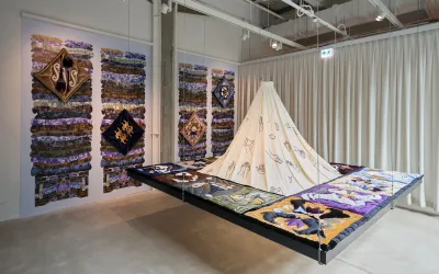 An installation suspended from the ceiling made of various textile pieces. Behind four textile pieces hang on the wall.