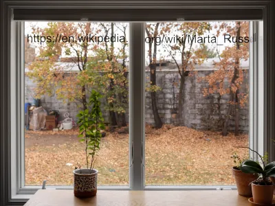 The web address of a wikipedia page on Marta Russell adhered to the top of a window looking out onto a fall yard scene. Potted house plants on an empty table sit in front of the window.