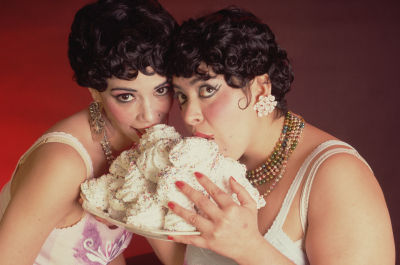 Two women licking a desserts piled on a plate.