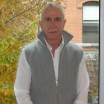 A man in a grey vest seated before a window looking out onto a tree.