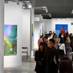 A crowded aisle at an art fair with various people looking at paintings in different booths.