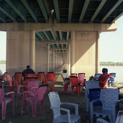 Several brightly colored chairs—in reds, pinks, and blues—are grouped together under a bridge overpass. A few people sit under them.