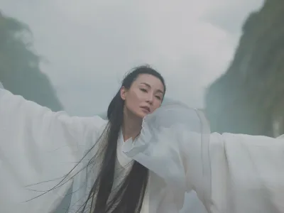 A woman in flowy fabric waving her arms in a mountainous landscape.
