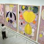 A woman staring at four large abstract paintings.
