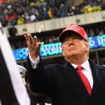 A man in a suit and a red hat tosses a coin at a stadium.