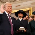 A suited man holds his right hand up next to two smartly dressed women.