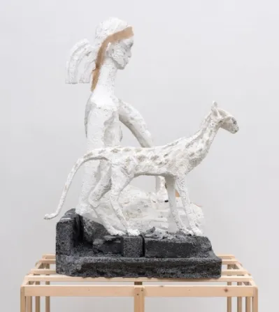 A styrofoam sculpture of a girl kneeling next to a slender cat atop of a wooden armature.