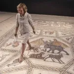US philantropist Shelby White poses for a picture on the roughly 1,700-year-old Lod mosaic, ahead of the inauguration of a new visitors centre she helped fund to display the prized piece in the mixed city of Lod in the centre of Israel, on June 27, 2022. - The Lod Mosaic Archaeological Center was specifically built to house the Roman mosaic dating back to the 3rd or 4th century CE, which was unearthed in the city in 1996 and described as 