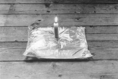 A black-and-white photograph of a lit candle in a pillow on a wood floor.