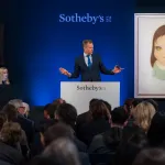 A man stands behind a rostrum with paintings either side of him.