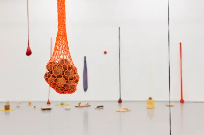 A sculpture resembling a pile of rocks suspended in hanging orange netting. Other similar works hang behind it.
