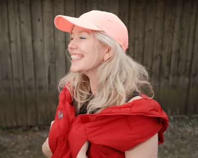 A smiling white woman in a pink baseball cap with her hands in her red vest