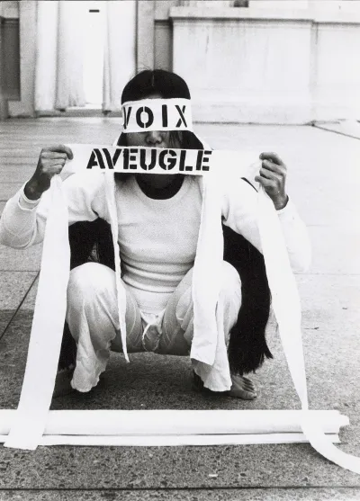 A woman dressed all in white is crouched down on a sidewalk. Her eyes and mouth are covered with a white piece of cloth that reads