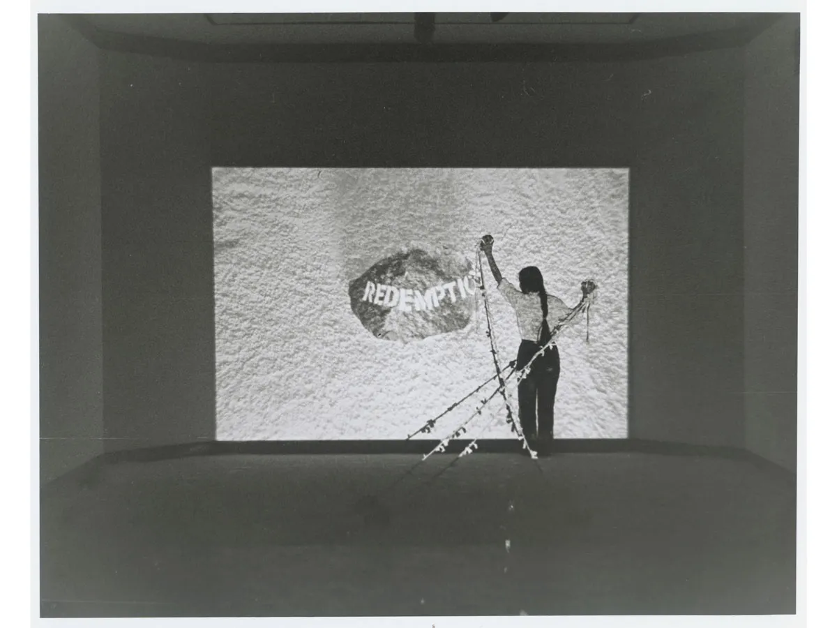 An archival photograph showing a woman holding three strings in front of a film projection which reads