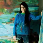 A white woman in a paint-spattered blue sweater placing one hand on an abstract canvas, with another canvas behind her.