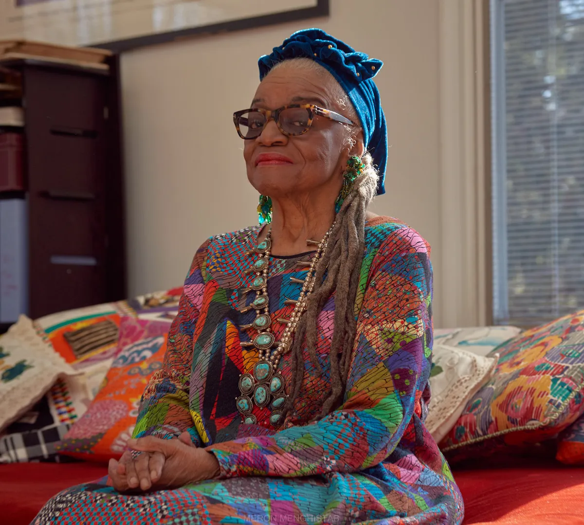 A Black woman in a blue turban and a multicolored shirt seated on a couch with patterned cushions.