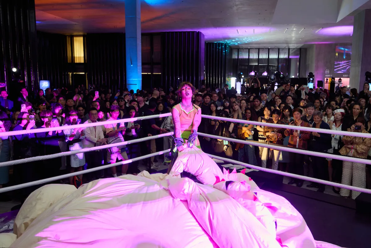 A performance artist wrestles a giant stuffed dragon in a wrestling ring in front of a crowd.