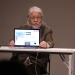 An elderly Asian man holding up a laptop screen to an audience.