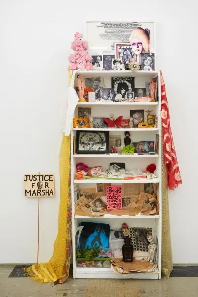 An altar-like sculpture in a white bookcase featuring various dolls, photographs, beads, and more.