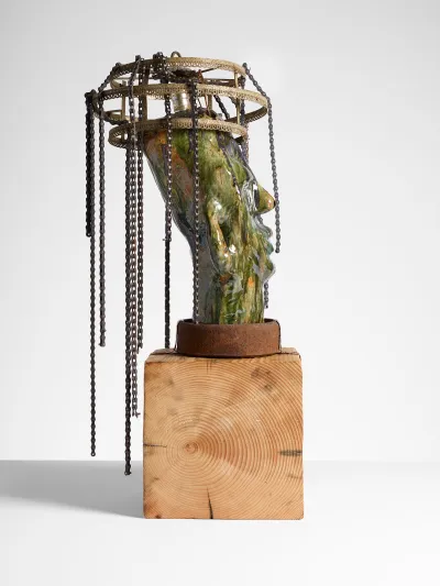 A sculpture of green-glazed ceramic head with a chandelier-like structure with chains on its head. It sits on a wooden base.