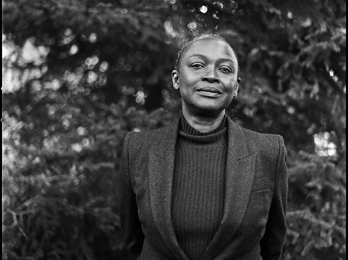 A black-and-white photograph of a Black woman in a vest standing amid trees.