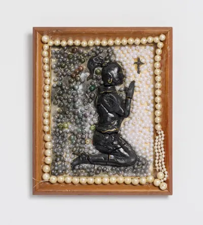 A mixed-media work, consisting of air dry clay, wood, Mod Podge, epoxy, acrylic, costume pearls, mother’s broken pearl necklace, showing a Black figure kneeling and praying to a small cross.