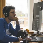 a man with headphones and blue sweater sits at a computer.