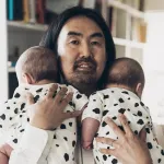A Japanese man with long hair holds twin babies.