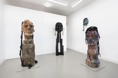 View of three large sculptures made of mixed media showing faces. One hangs at the wall behind.