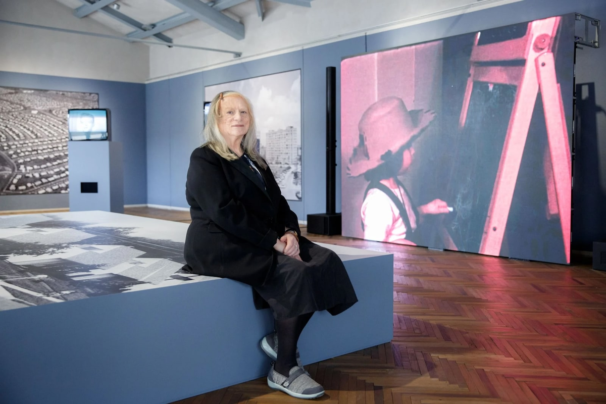A woman seated on a platform in front of video monitors.