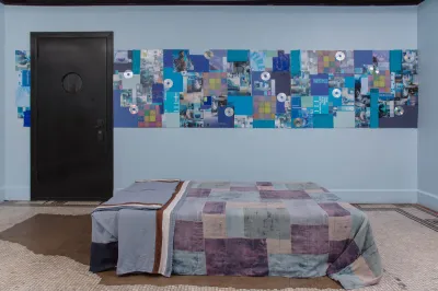 Installation view of a table covered with a quilt to look like a bed. Several blue paintings with CDs hang on a light-blue wall behind.