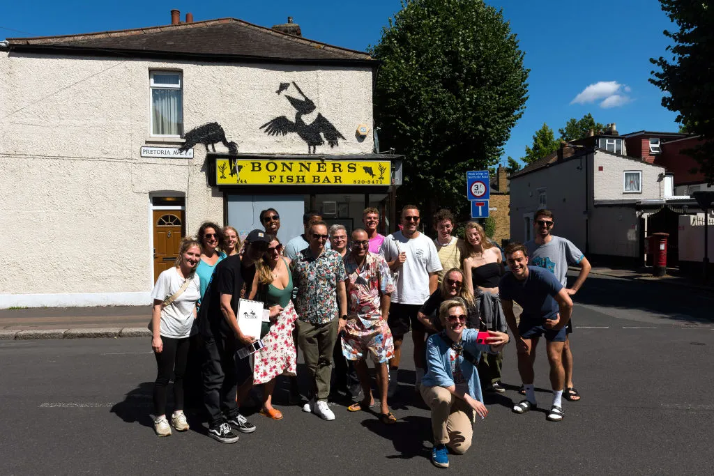 WALTHAMSTOW, ENGLAND - AUGUST 09: A group of people take a selfie in front of mural purportedly by the artist Banksy, showing pelicans eating fish, on the side of Bonners fish and chip shop on August 09, 2024 in Walthamstow, England. It was the fifth artwork debuted by the elusive street artist in London this week. (Photo by Matthew Baker/Getty Images)