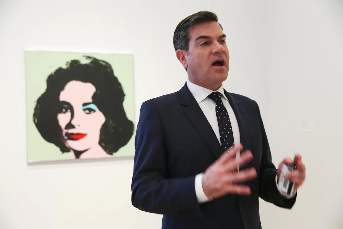 A man in a suit talking in front of a painting.