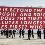 A group of soldiers beneath a red mural with text that begins 