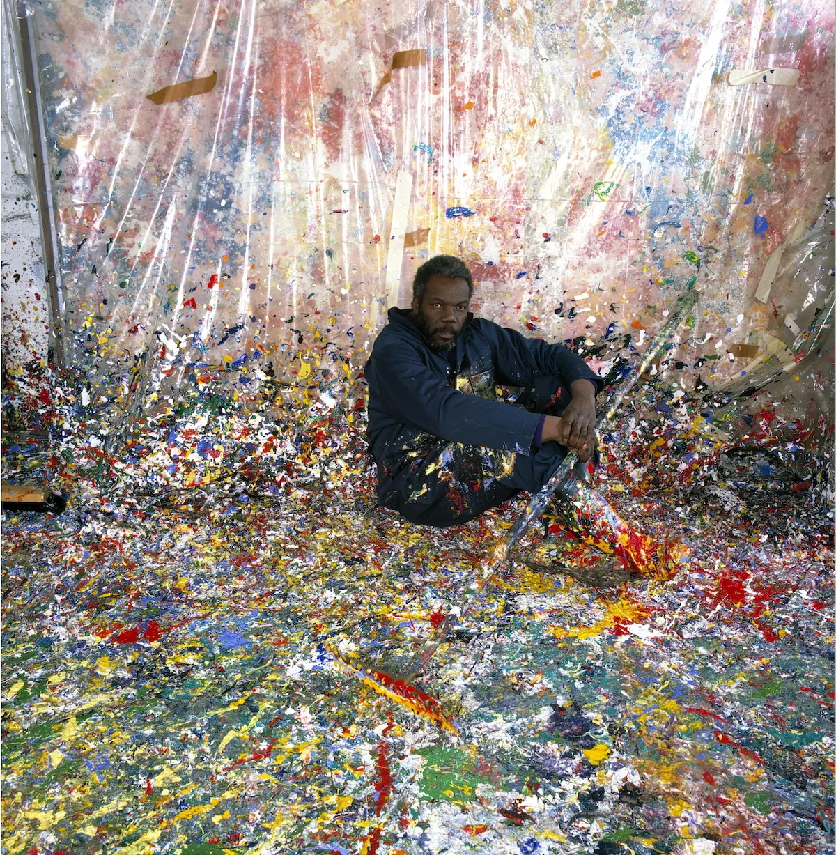 A painting of a man seated in a studio whose floor is covered in paint.
