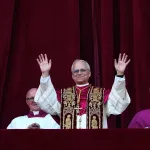 A close-up photo of a balcony with dark red drapes. A man in white robes standing, smiling, with his hands raised. Three other men are seated slightly behind him.