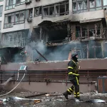 A burning building with smoke and a man in a black and yellow protective suit.