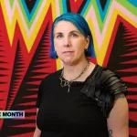 A woman with dyed-blue hair standing in front of a weaving featuring zigzagging lines in various colors.