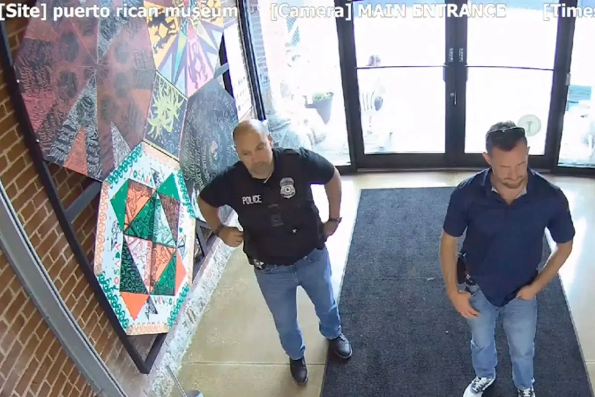 Two men stand in a lobby as seen by security footage. One wears a vest that says police and has a badge. The other does not.