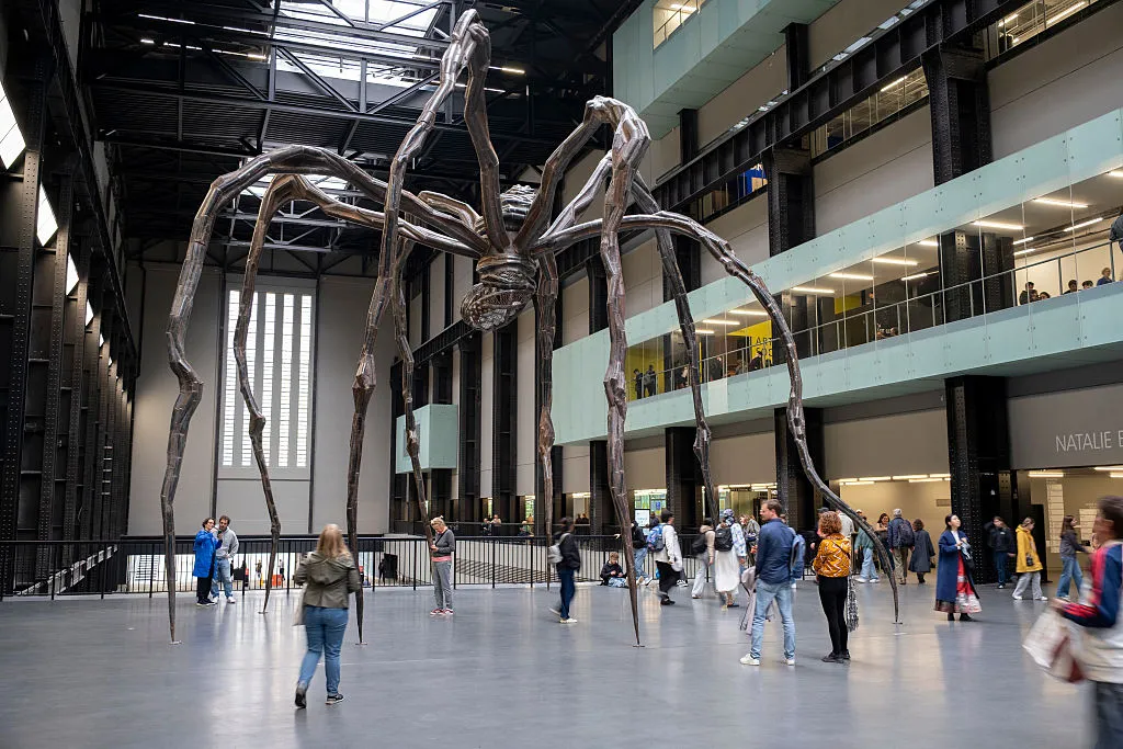 A sculpture of a giant spider in a big room with people walking underneath it.