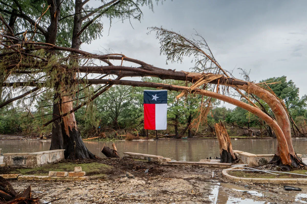 CENTER POINT, TEXAS - JULY 13: A Texas flag hangs from a storm-damaged tree on the banks of the Guadalupe River on July 13, 2025 in Center Point, Texas. More than 160 people are still missing after storm cells halted over the area, dumping nearly 15 inches of rain and causing a 22-foot rise along the Guadalupe River. (Photo by Brandon Bell/Getty Images)